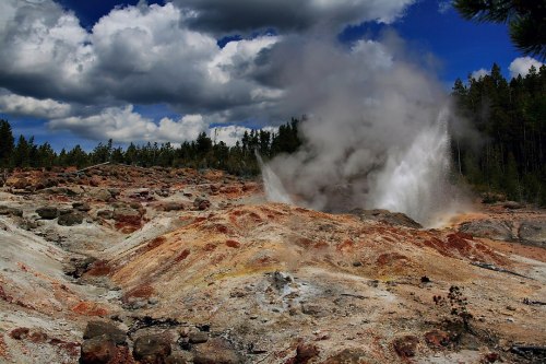 A geyser erupts amidst a rocky landscape, surrounded by trees and dramatic clouds in the sky.