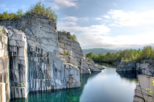 A serene quarry landscape with steep stone cliffs and a calm, turquoise lake surrounded by greenery under a cloudy sky.