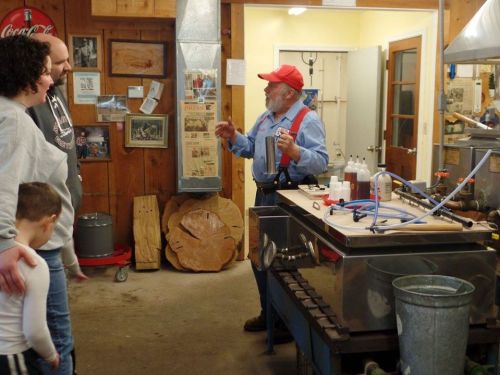 A man in a red cap explains something to a family in a rustic kitchen with wooden walls and equipment.