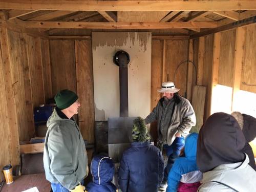 A group of people listens to a speaker in a wooden shed with a stove, discussing a topic related to the space.