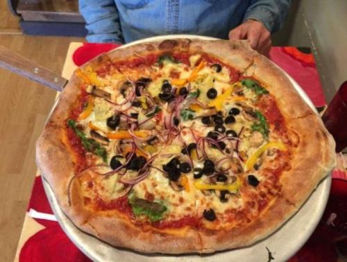 A large pizza topped with vegetables, olives, and cheese on a silver platter, with a person’s hands visible in the background.