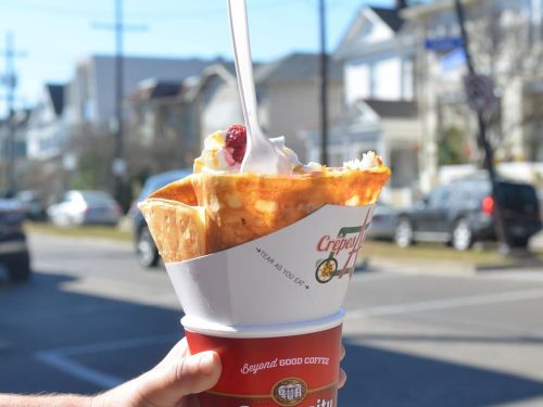 A hand holds a large crepe filled with whipped cream and a cherry, with a street scene in the background.