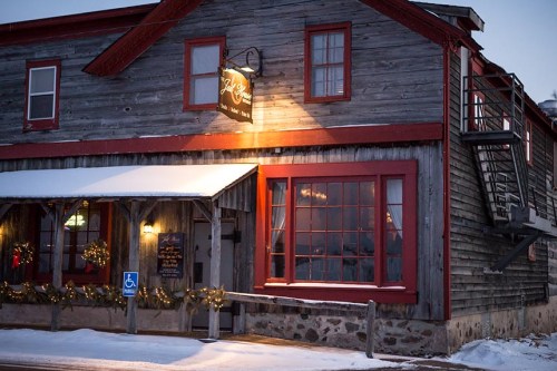 A rustic wooden building with red accents, snow-covered, featuring a warm light and holiday decorations.