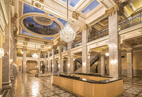 Elegant lobby with marble columns, ornate chandeliers, and a painted ceiling, featuring intricate tile flooring.