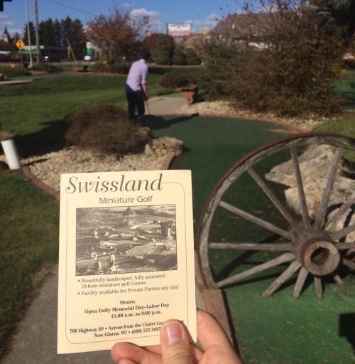 A person plays mini golf at Swissland, with a flyer detailing hours and features in the foreground.