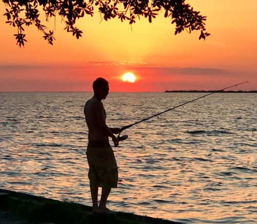 A silhouette of a person fishing at sunset, with vibrant orange and pink hues reflecting on the water.