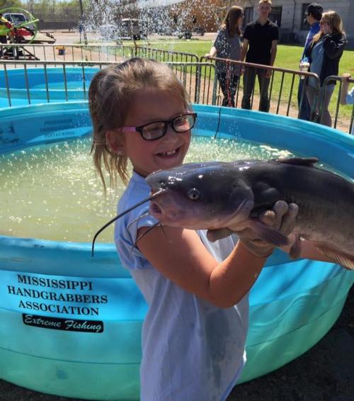 A girl in glasses proudly holds a large catfish beside a blue fishing tank at an outdoor event.