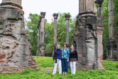 Three women stand smiling among ancient ruins with tall columns, surrounded by greenery.