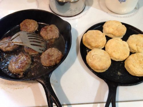 Two cast iron skillets: one with sizzling sausage patties and the other with freshly baked biscuits.