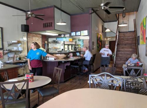 A cozy café interior with patrons at tables, a counter, and a staircase, featuring colorful artwork on the walls.
