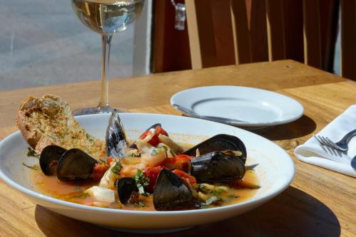 A plate of seafood stew with mussels, vegetables, and a slice of bread, accompanied by a glass of white wine.
