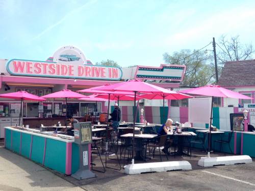A colorful drive-in restaurant with pink umbrellas, outdoor seating, and customers enjoying their meals on a sunny day.