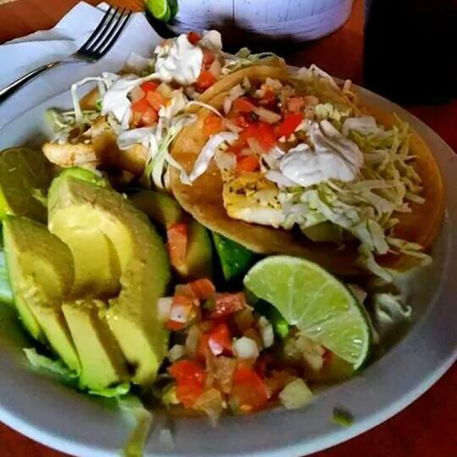 A plate of tacos topped with lettuce, tomatoes, and sour cream, served with avocado slices and lime wedges.