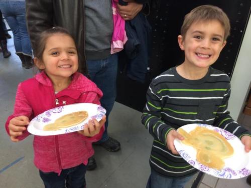 Two smiling children hold paper plates with pancake faces, standing in a casual indoor setting.