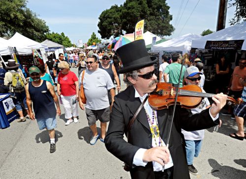 A street festival scene with a crowd walking by, while a man in a top hat plays the violin. Colorful tents line the street.