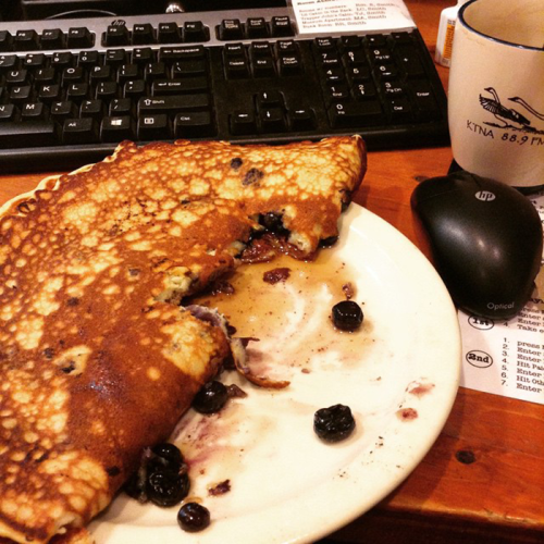 A plate with a half-eaten blueberry pancake, a keyboard, a mouse, and a coffee mug in the background.
