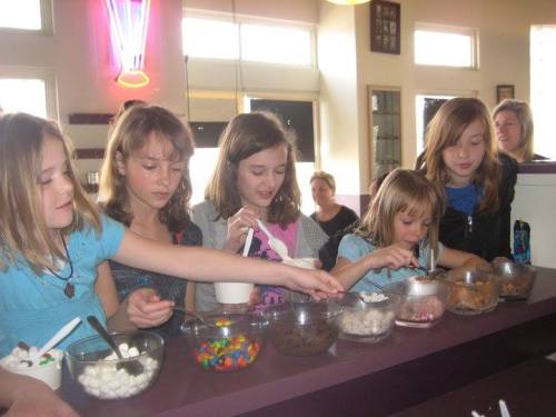 A group of five girls enjoying a dessert bar, selecting toppings from bowls filled with candies and marshmallows.