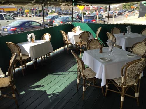 Outdoor dining area with white tablecloths and wicker chairs, surrounded by green curtains and a view of parked cars.