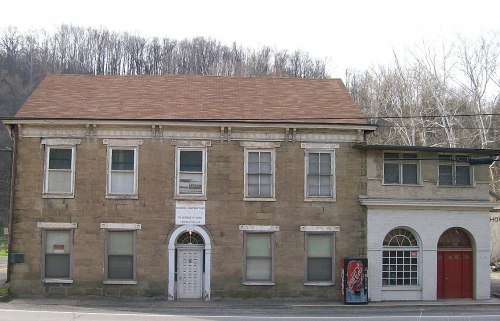 Historic stone building with a brown roof, featuring multiple windows and a sign, set against a wooded hillside.