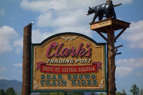Sign for Clark's Trading Post featuring bear shows and train rides, with a bear statue on top against a blue sky.