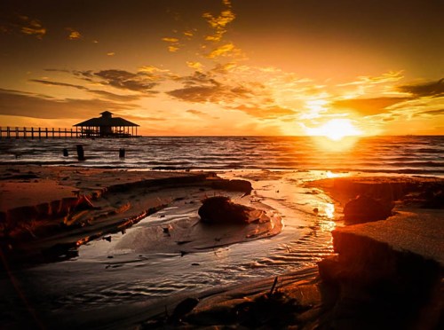 Sunset over a calm sea with a pier and a small gazebo silhouetted against the vibrant orange and yellow sky.