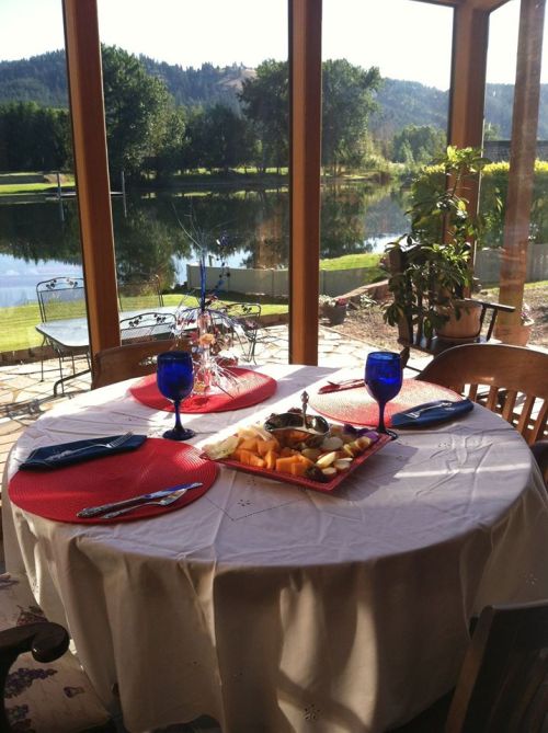 A beautifully set dining table with blue glasses and a fruit platter, overlooking a serene lake and lush greenery.