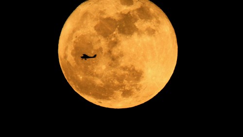 A silhouette of an airplane flying in front of a large, glowing orange full moon against a dark sky.