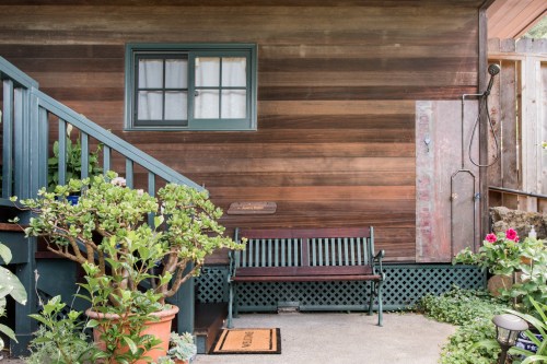 A wooden bench sits beside a plant-filled patio, with a shower and window on a rustic wall.
