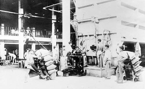 Black and white photo of workers in a factory, operating machinery and handling large bags.