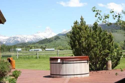 A wooden hot tub on a deck with a scenic view of snow-capped mountains and green fields in the background.