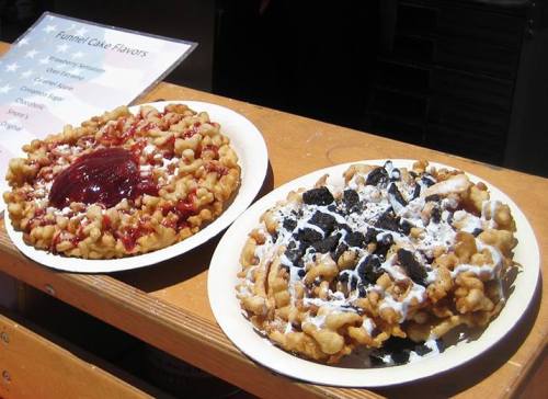 Two plates of funnel cakes topped with strawberry sauce and crushed cookies, drizzled with white icing.