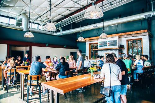 A lively restaurant interior with people dining and socializing at wooden tables under industrial-style lighting.