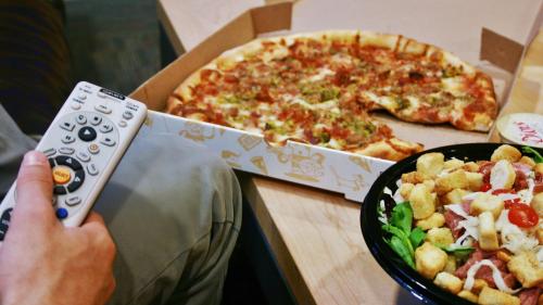 A hand holding a remote control next to a pizza box and a bowl of salad on a table.