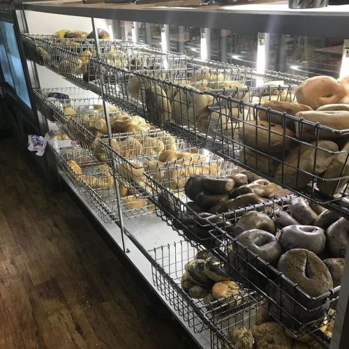 A display case filled with various types of bagels and baked goods on wire racks, illuminated from above.