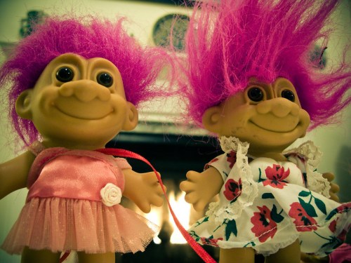 Two colorful troll dolls with pink hair, one in a pink dress and the other in a floral dress, smiling in front of a fireplace.