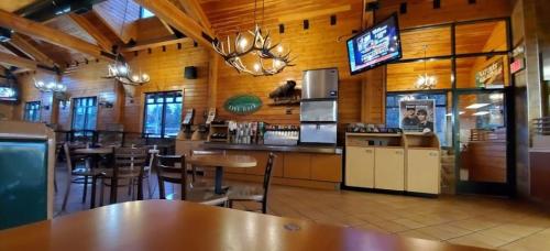 Interior of a rustic restaurant with wooden beams, tables, and a beverage station, featuring large windows and a TV.