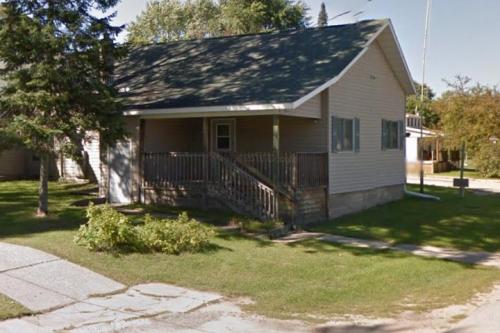 A small, light-colored house with a sloped roof, front porch, and surrounding greenery on a sunny day.