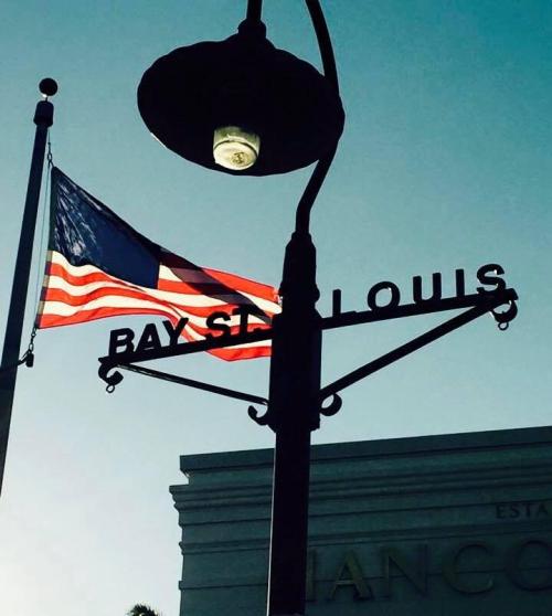 A street sign for Bay St. Louis with an American flag waving in the background against a clear sky.