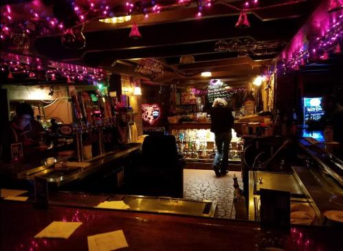 A dimly lit bar with colorful neon lights, shelves of liquor, and a person walking towards the back.