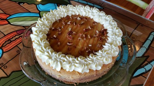 A round cheesecake topped with whipped cream, caramel drizzle, and chopped pecans, displayed on a decorative plate.