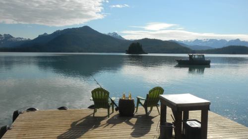 A serene lakeside view with two green chairs, a table, and a boat on calm waters surrounded by mountains.