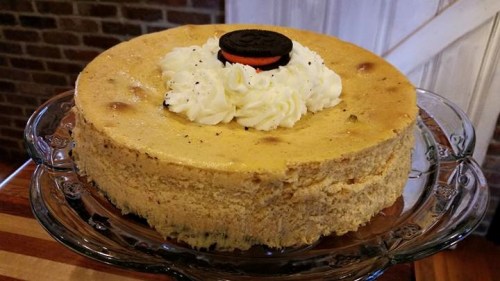 A creamy cheesecake topped with whipped cream and an Oreo cookie, displayed on a glass cake stand.