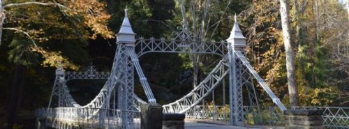 A decorative metal suspension bridge surrounded by trees with autumn foliage.