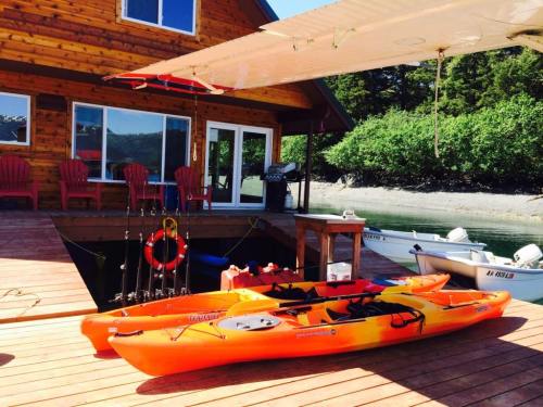 Two orange kayaks are docked on a wooden deck beside a house, with fishing gear and a grill in the background.