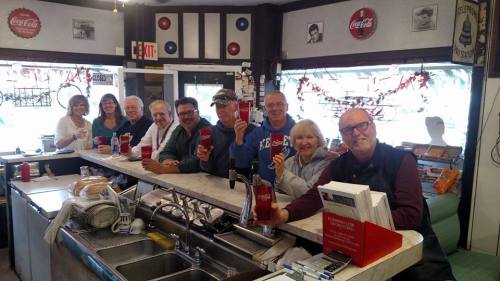A group of people smiling and holding drinks at a diner counter, with retro decor and a cozy atmosphere.