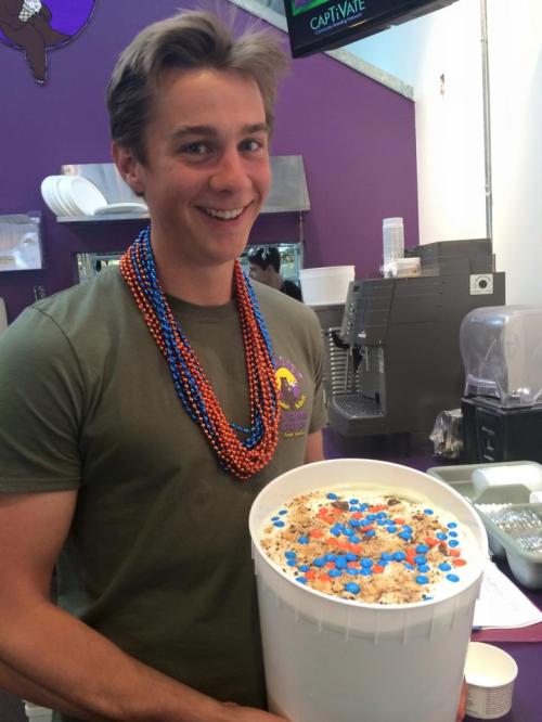 A smiling young man wearing a colorful bead necklace holds a large bucket of ice cream in a shop.