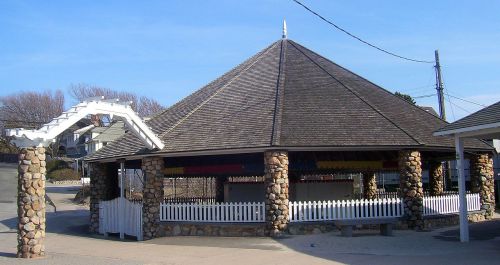 A stone pavilion with a wooden roof, surrounded by a white picket fence, set against a clear blue sky.