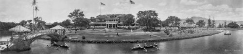 A panoramic view of a historic building by a river, featuring boats, a bridge, and lush greenery.