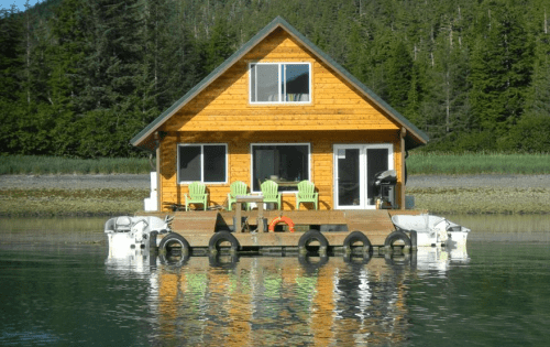 A wooden cabin on stilts floats on calm water, surrounded by lush greenery and featuring a deck with chairs.