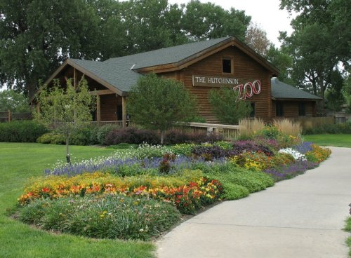 A landscaped path leads to the wooden entrance of the Hutchinson Zoo, surrounded by colorful flowers and greenery.
